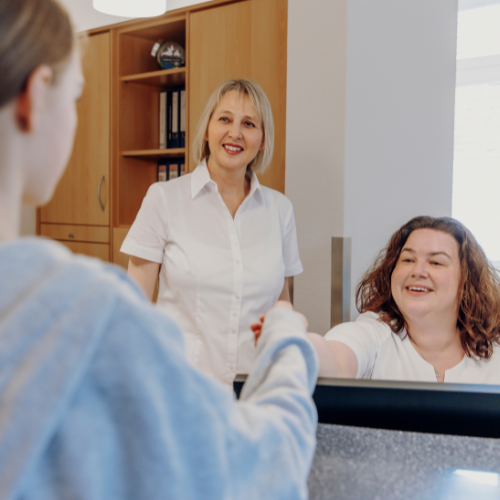 Ein freundlicher Empfangsbereich mit Frau Dr. Grüneisl und einer Mitarbeiterin in weißen Hemden, die eine Patientin in einem blauen Oberteil begrüßen. Dieses Bild vermittelt eine warme und einladende Atmosphäre der Praxis.